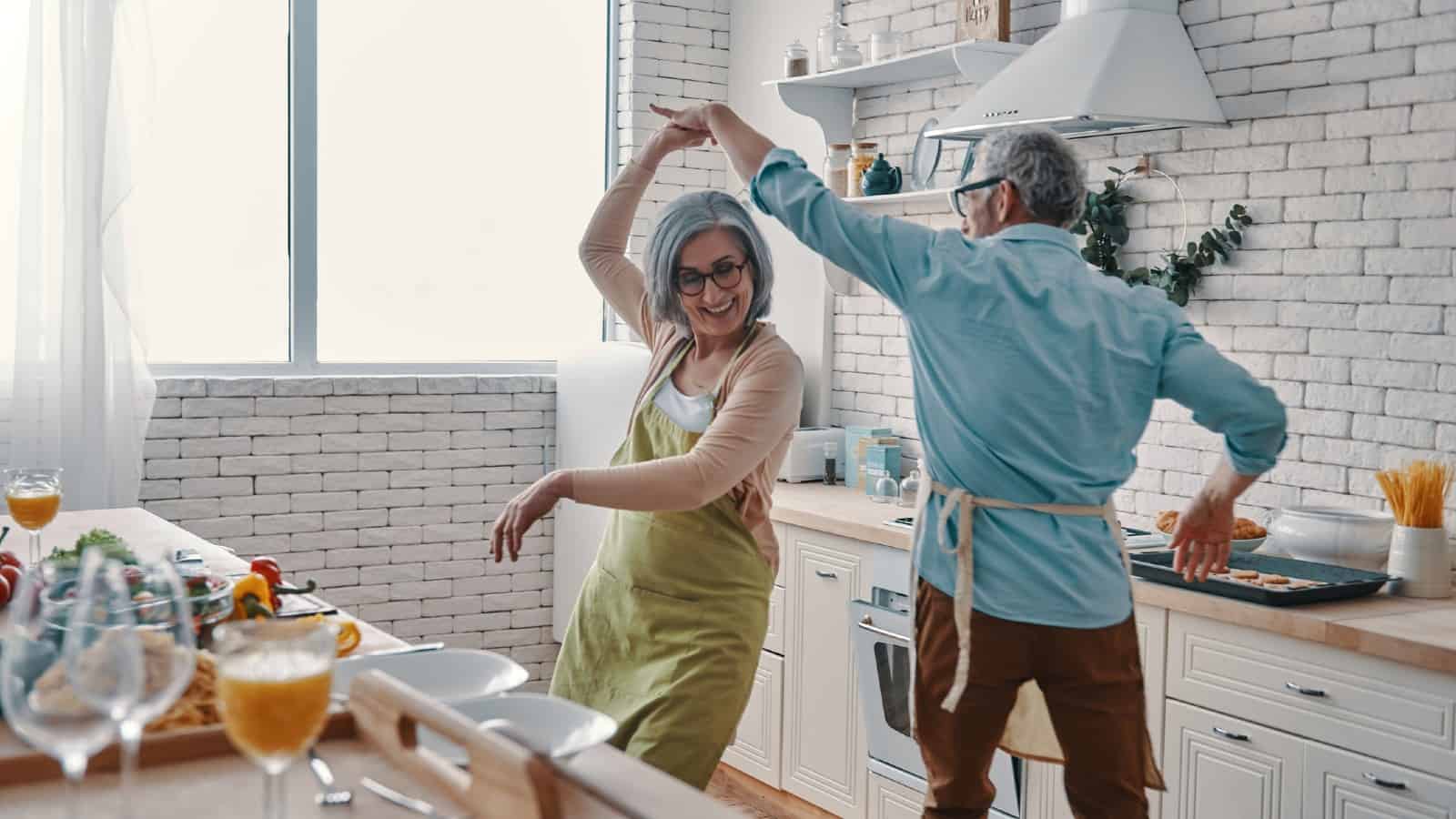 Παιχνιδιάρικο ηλικιωμένο ζευγάρι με ποδιές χορεύει και χαμογελά ενώ ετοιμάζει υγιεινό δείπνο στο σπίτι